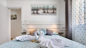 A hotel room with a bed, two towels shaped like swans, a painting of trees, and a window with curtains. - Vacation Home "Sol y Mar Caleta de Fuste" with Shared Pool, Garden & Wi-Fi (Caleta de Fuste)