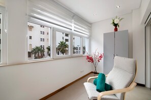 Une pièce avec une fenêtre, une chaise, un meuble et un vase rempli de fleurs.
