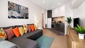 Un salon moderne avec un canapé, des coussins colorés, un tableau et une cuisine en arrière-plan.