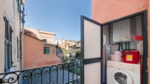 Ein Balkon mit einer Waschmaschine, einem roten Eimer und einem weißen Behälter.