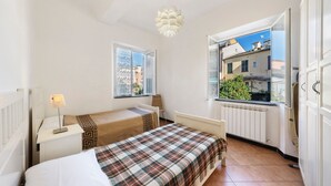 Ein Schlafzimmer mit einem Bett, einem Nachttisch, einer Lampe, einem Fenster mit Blick auf Gebäude und einer Heizung.