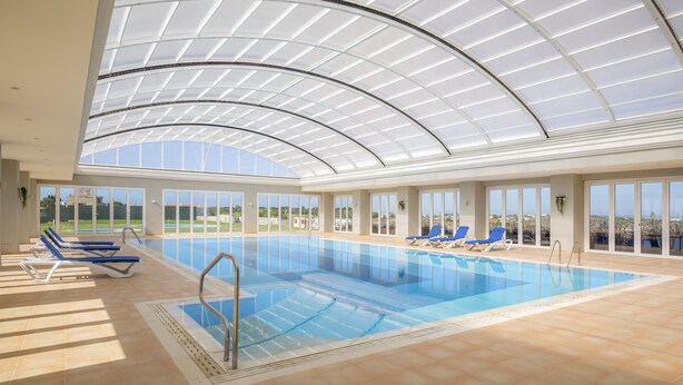 An indoor swimming pool with a glass ceiling, blue lounge chairs, and a view of the landscape. - Casa da Boa Vida at Boa Vista Golf Resort, Bayview Village (Lagos)