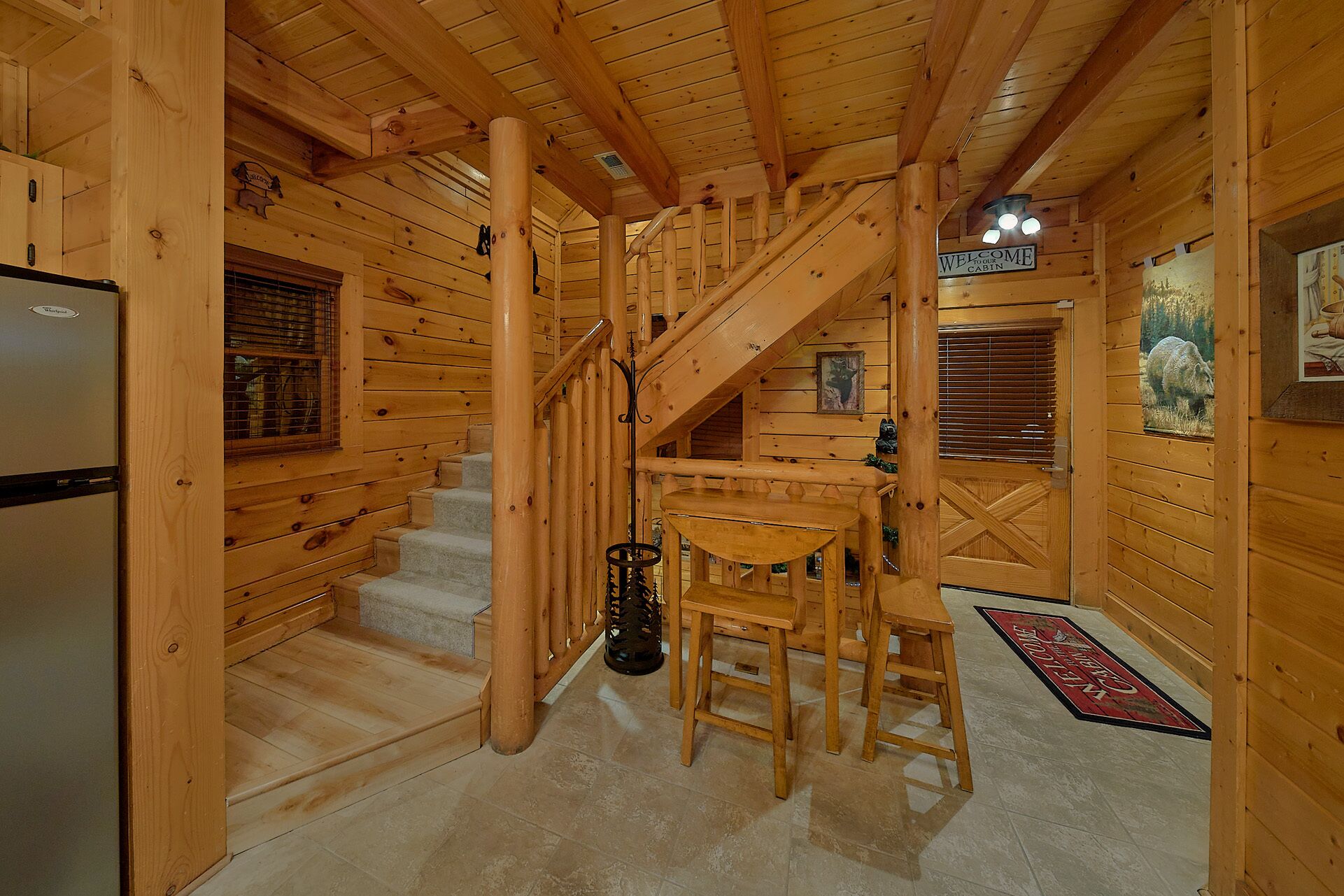 A wooden cabin interior with a staircase, a dining area with a table and chairs, and a refrigerator.