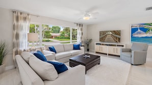 A modern living room with a sofa, coffee table, and armchairs. There is a large window with curtains, a ceiling fan, and a painting on the wall. - Luxurious Pool Home Central to Everything! (Palm Beach Gardens)