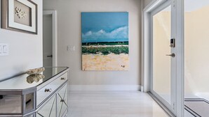 A hallway with a glass door leading to a balcony, a framed artwork on the wall, and a reflective surface on a piece of furniture. - Luxurious Pool Home Central to Everything! (Palm Beach Gardens)