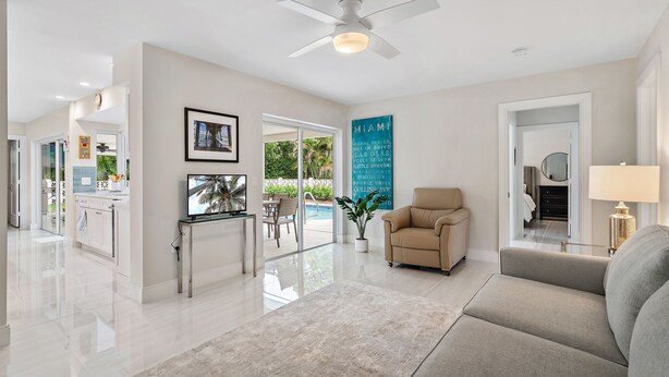 A modern living room with a sofa, a coffee table, and a TV. There is a ceiling fan, a door leading to another room, and a window with a view of the outdoors. - Luxurious Pool Home Central to Everything! (Palm Beach Gardens)