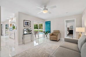 A modern living room with a sofa, a coffee table, and a TV. There is a ceiling fan, a door leading to another room, and a window with a view of the outdoors. - Luxurious Pool Home Central to Everything! (Palm Beach Gardens)
