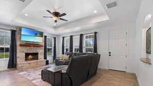 A spacious living room with a fireplace, a large flat-screen TV, a ceiling fan, and a view of the outdoors through large windows.
