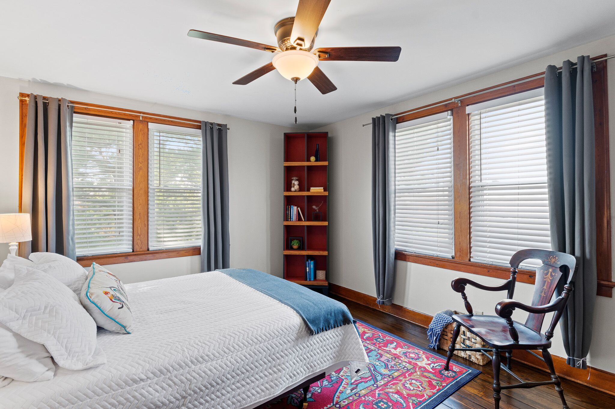 Une chambre à coucher comprenant un lit, une chaise, un ventilateur de plafond, une bibliothèque et de grandes fenêtres équipées de stores.