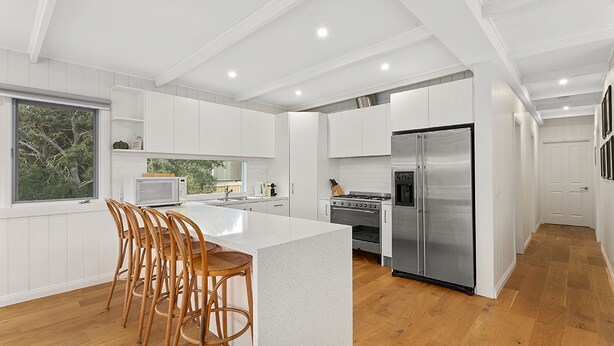 Una cocina moderna con armarios blancos, una isla grande con taburetes, electrodomésticos de acero inoxidable y vistas a través de un ventanal.