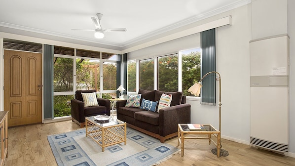 A living room with a brown sofa, two armchairs, a coffee table, and a floor lamp. There are large windows with a view of trees and a door with a window.