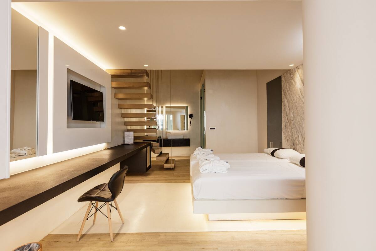Una habitación de hotel moderna con una cama grande, un escritorio con silla, un televisor de pantalla plana en la pared y una escalera que conduce a otro nivel.