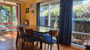 A dining area with a round table and chairs, large windows with curtains, and a view of greenery outside.