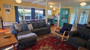 A living room with a sofa, armchairs, a coffee table, and a rug. There is a dining area with a table and chairs, and a kitchen with cabinets and a sink.