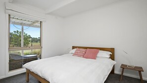 A bedroom with a large bed, a nightstand, a window with a view of greenery, and a small table.