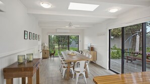 A spacious dining area with a long wooden table and white chairs, large windows, and a view of the outdoors.
