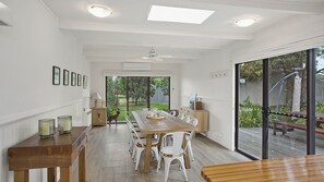 A spacious dining area with a long wooden table and white chairs, large windows, and a view of the outdoors.