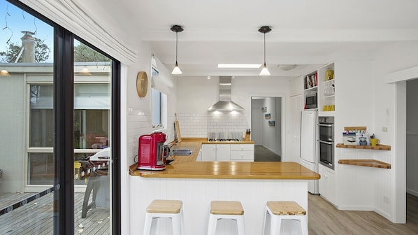A modern kitchen with white cabinets, wooden countertops, and a central island with stools. There are pendant lights, a stainless steel range hood, and a refrigerator.