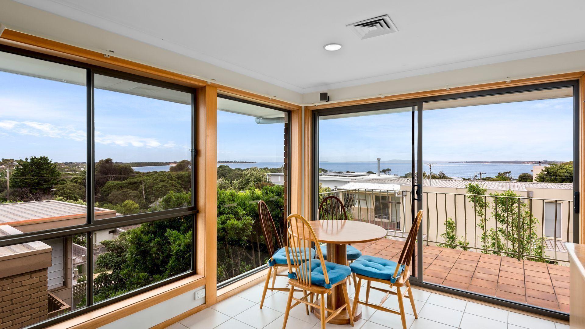 A balcony with a table and chairs, large windows with a view of the sea and greenery.