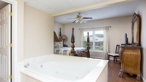 A bedroom with a large white bathtub, a ceiling fan, a wooden dresser, and a window with blinds. - Riverfront Condo Pigeon Forge/Mt. LeConte Views (Pigeon Forge)