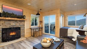 A cozy cabin interior with a stone fireplace, a large flat-screen TV, a dining area, and a sofa with a view of the outdoors. - Outstanding Mt. LeConte View/Indoor Pool & Hot Tub (Gatlinburg)