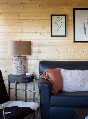 Una habitación con pared de madera, un sofá de cuero negro con una manta blanca mullida, una lámpara sobre un pedestal de madera e impresiones botánicas enmarcadas.