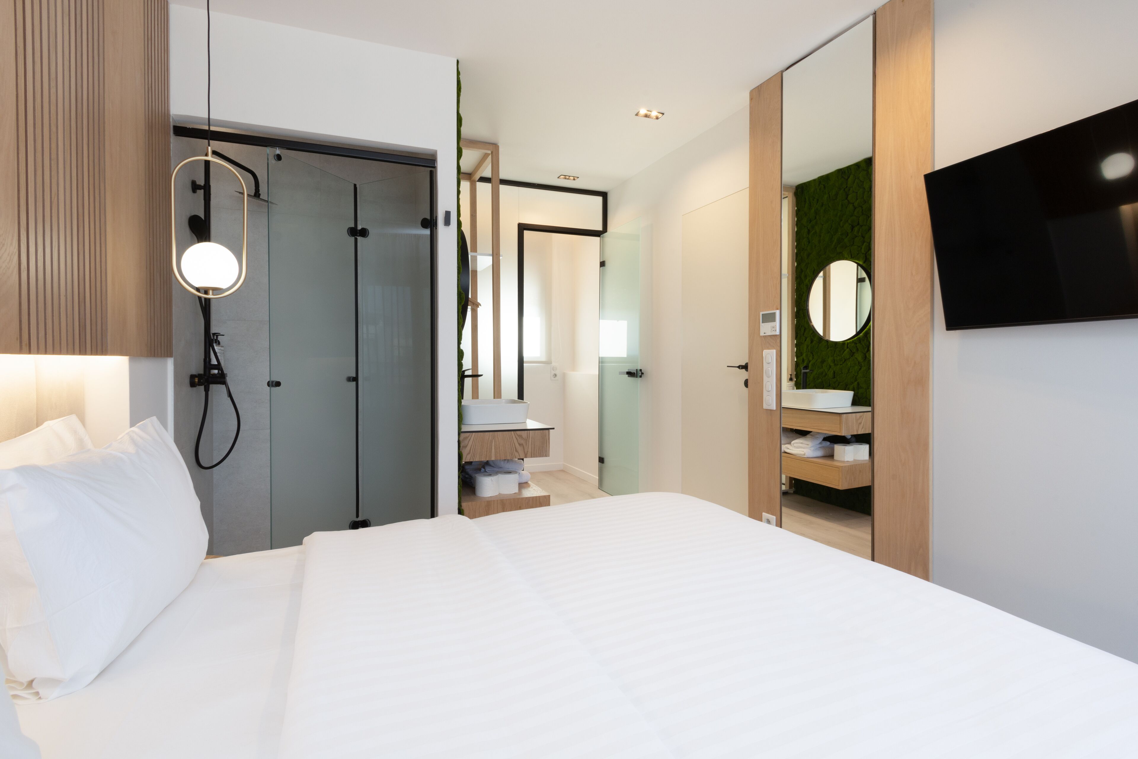 Une chambre d’hôtel moderne dotée d’un grand lit, d’un dressing, d’une salle de bain avec un miroir et un lavabo, et d’un téléviseur à écran plat fixé au mur.