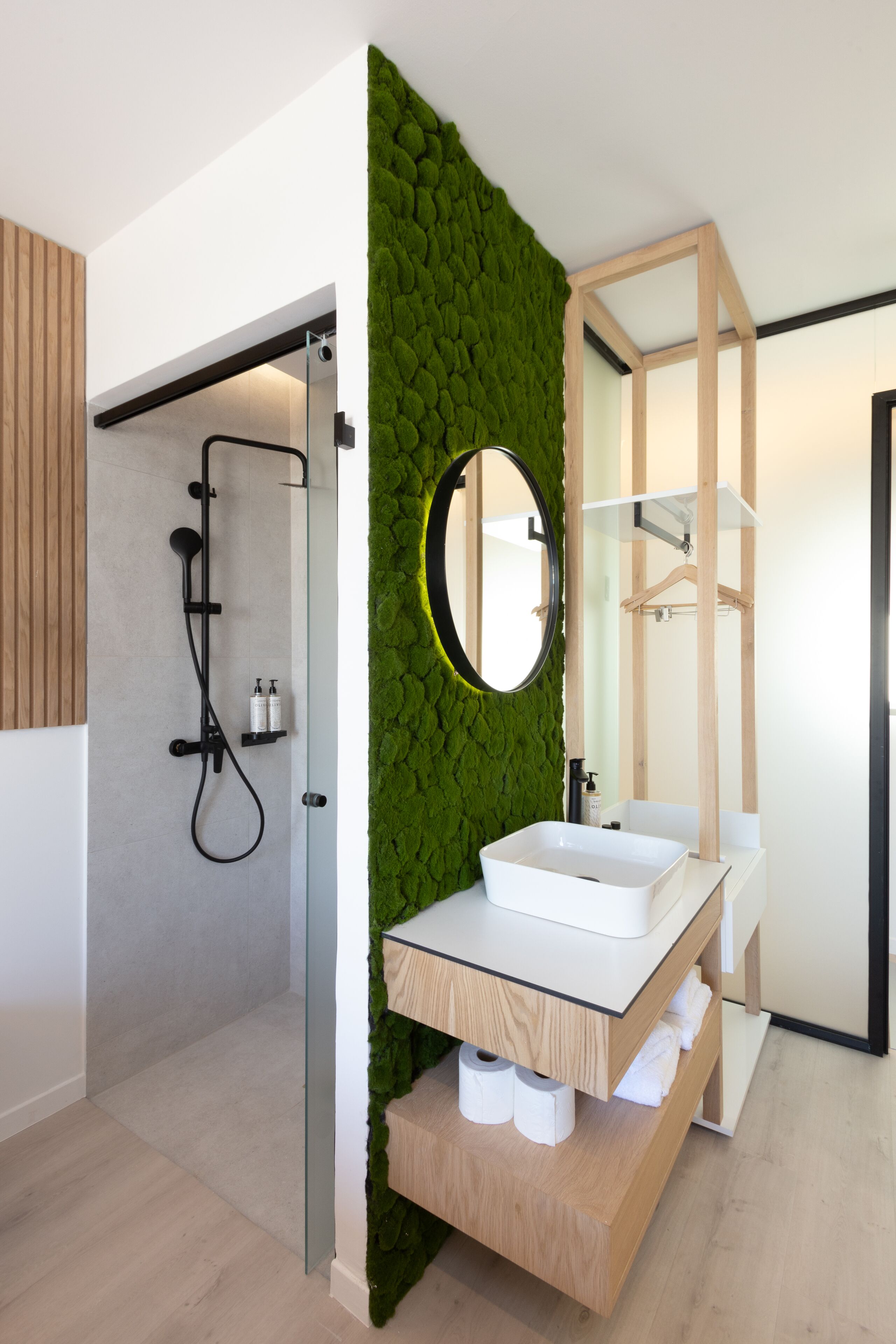 A modern bathroom with a green textured wall, a wooden vanity, a white sink, and a glass-enclosed shower area.
