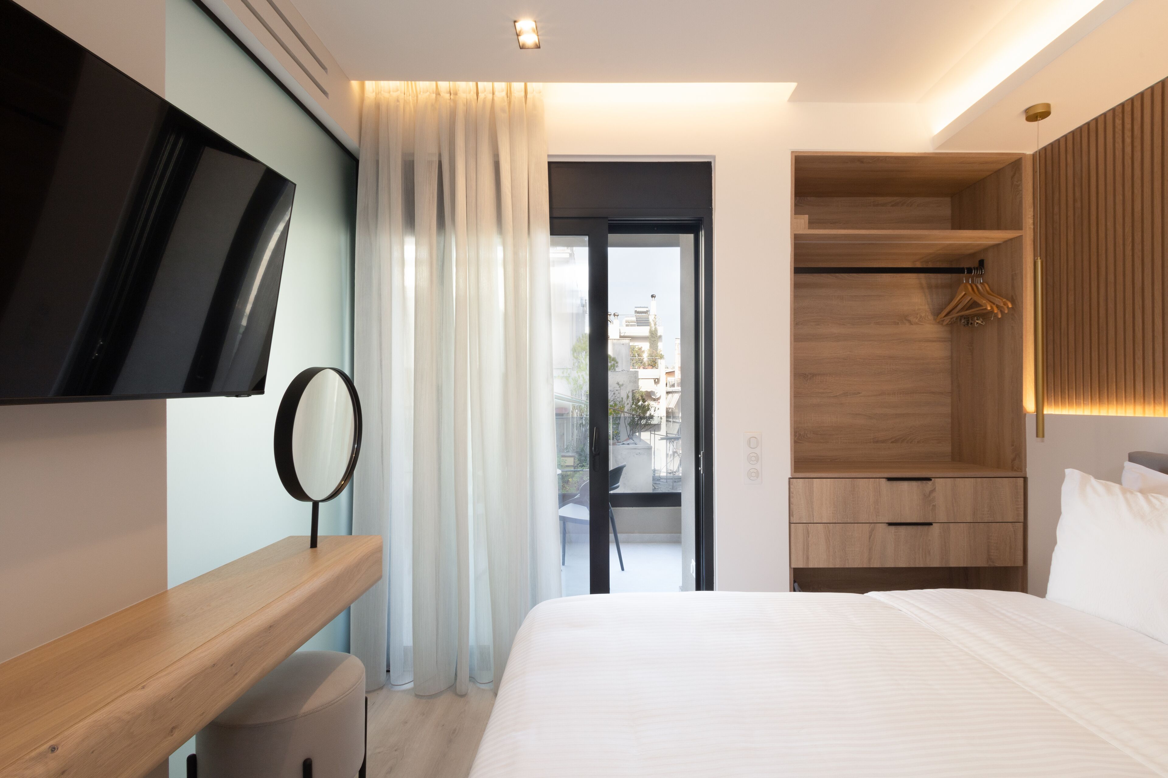 Una habitación de hotel moderna con una cama grande, un televisor de pantalla plana en la pared, una mesita de noche de madera, un espejo y un armario con perchero.