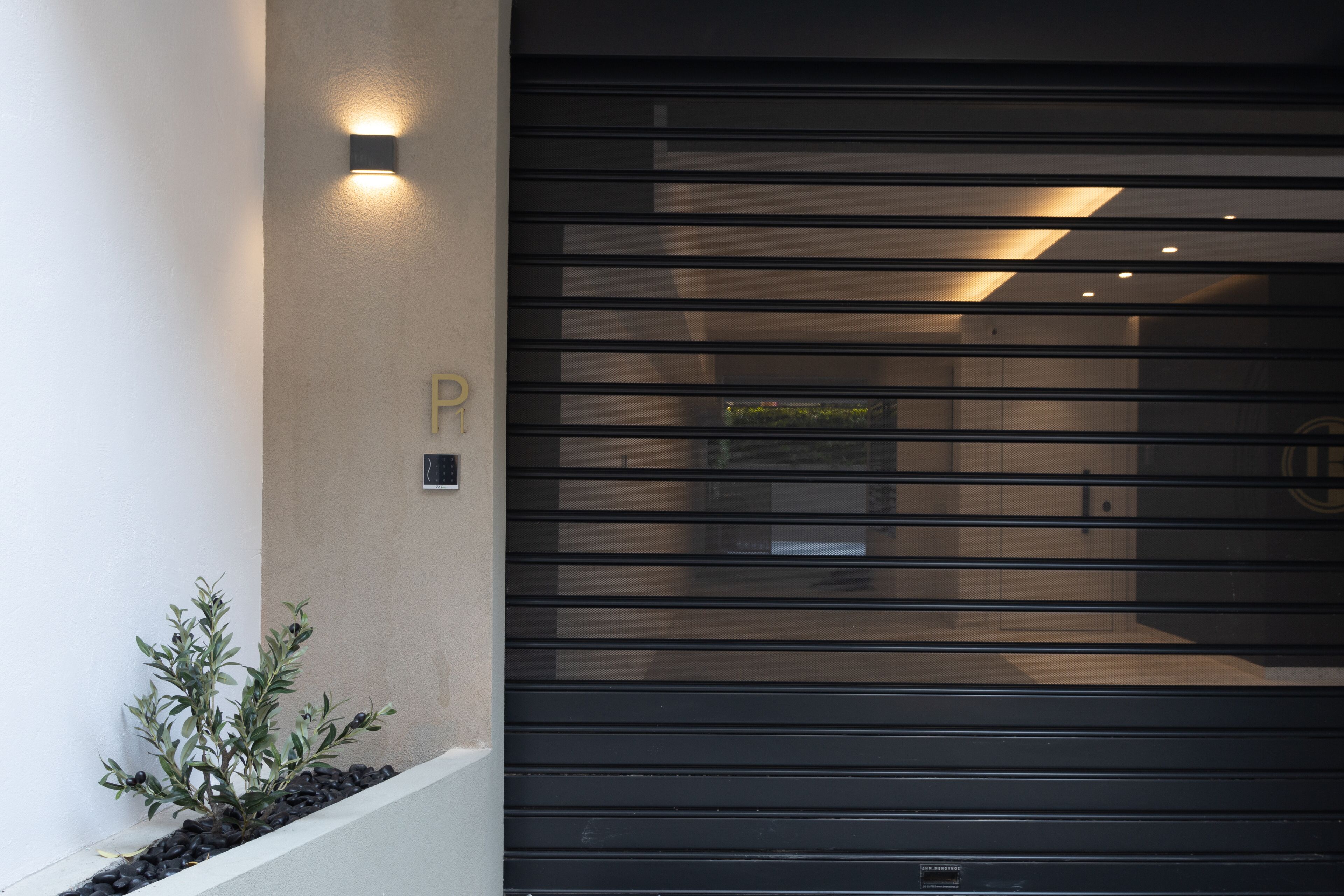 Entrada d'un edifici modern amb una persiana metàl·lica tancada, una petita planta en test i un llum penjant a la paret.