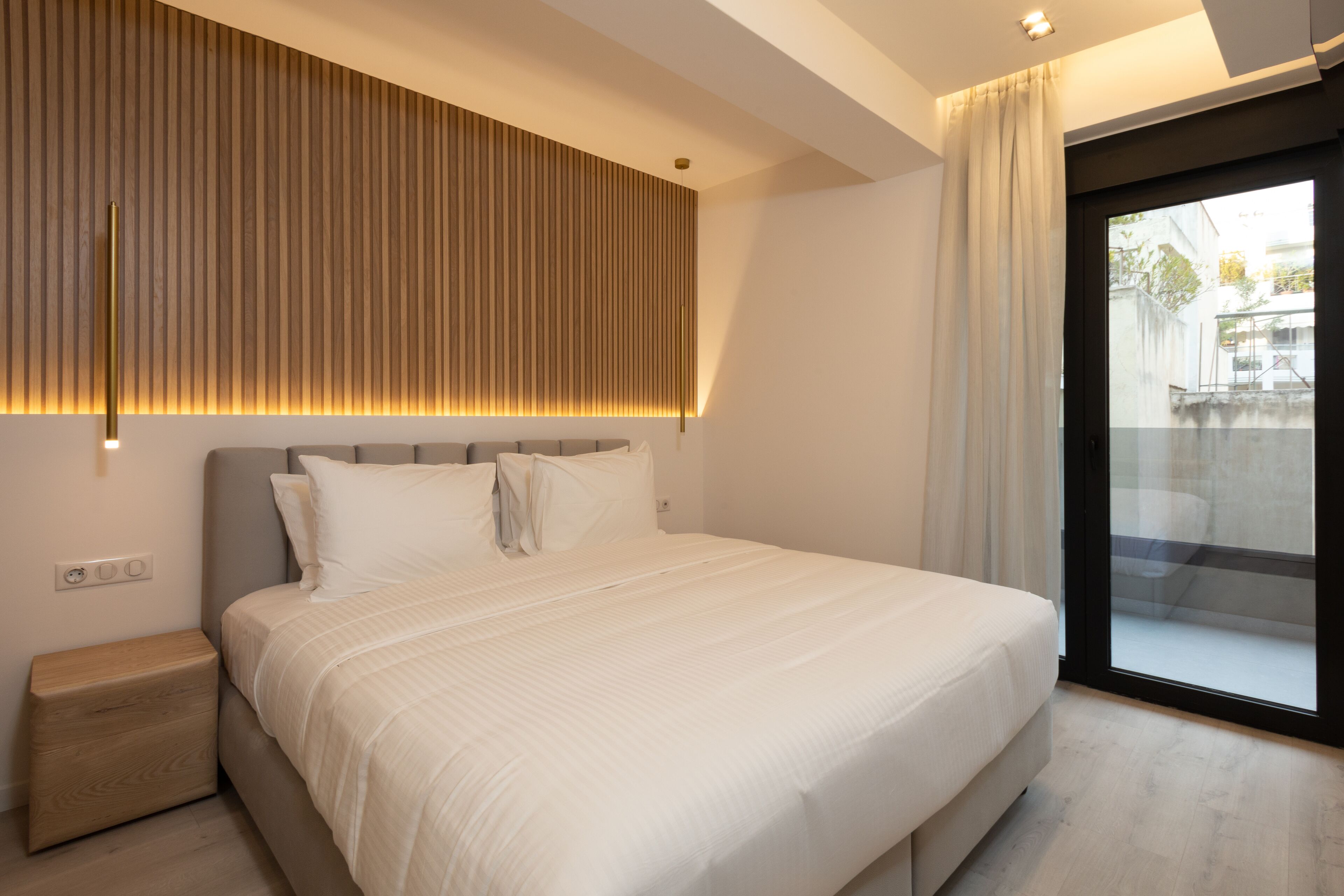 A modern bedroom with a large bed, a bedside table, a wall-mounted light, and a sliding glass door to the outside.