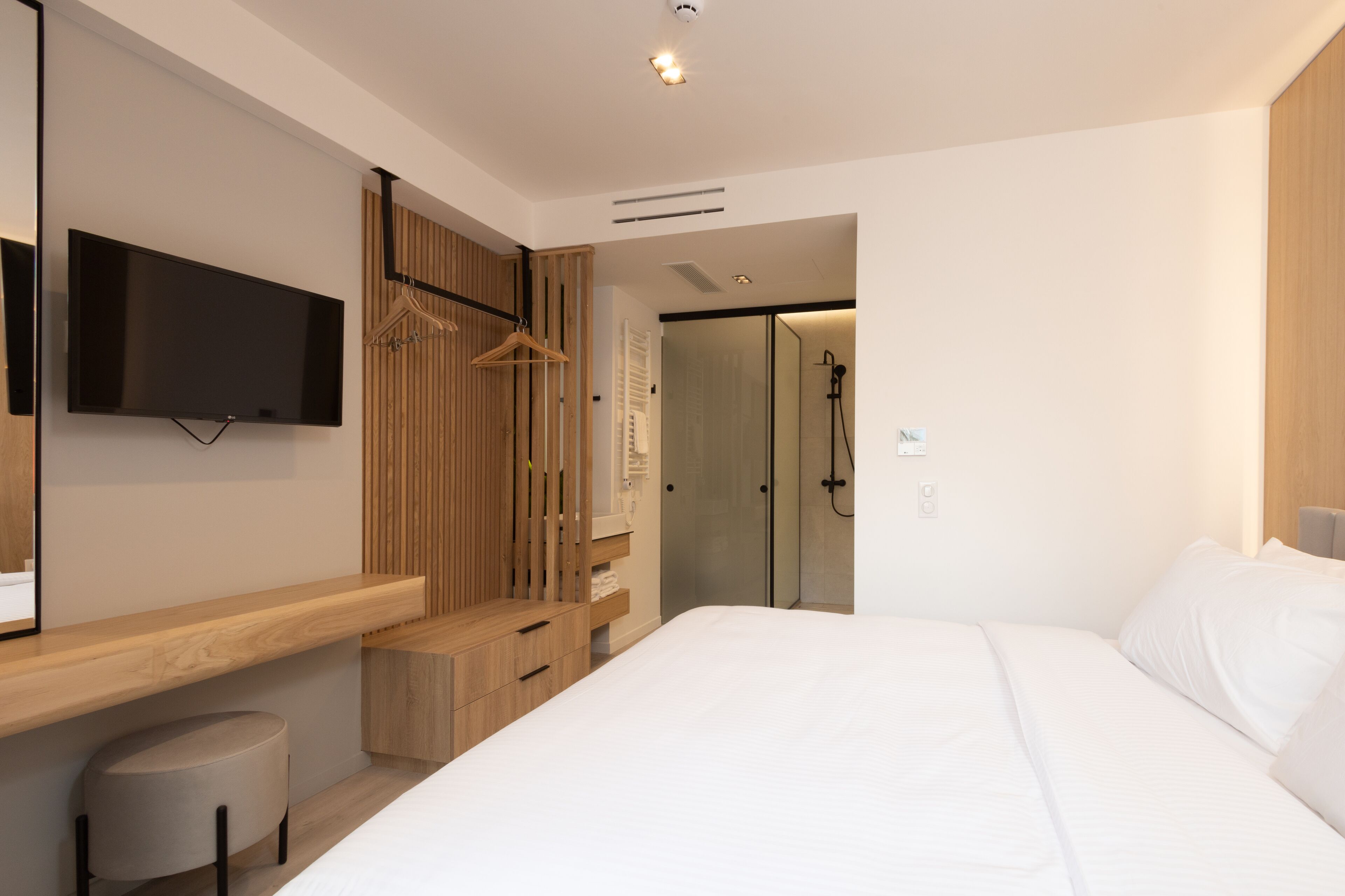 A modern bedroom with a large bed, a TV mounted on the wall, a wooden wardrobe, and a walk-in closet.