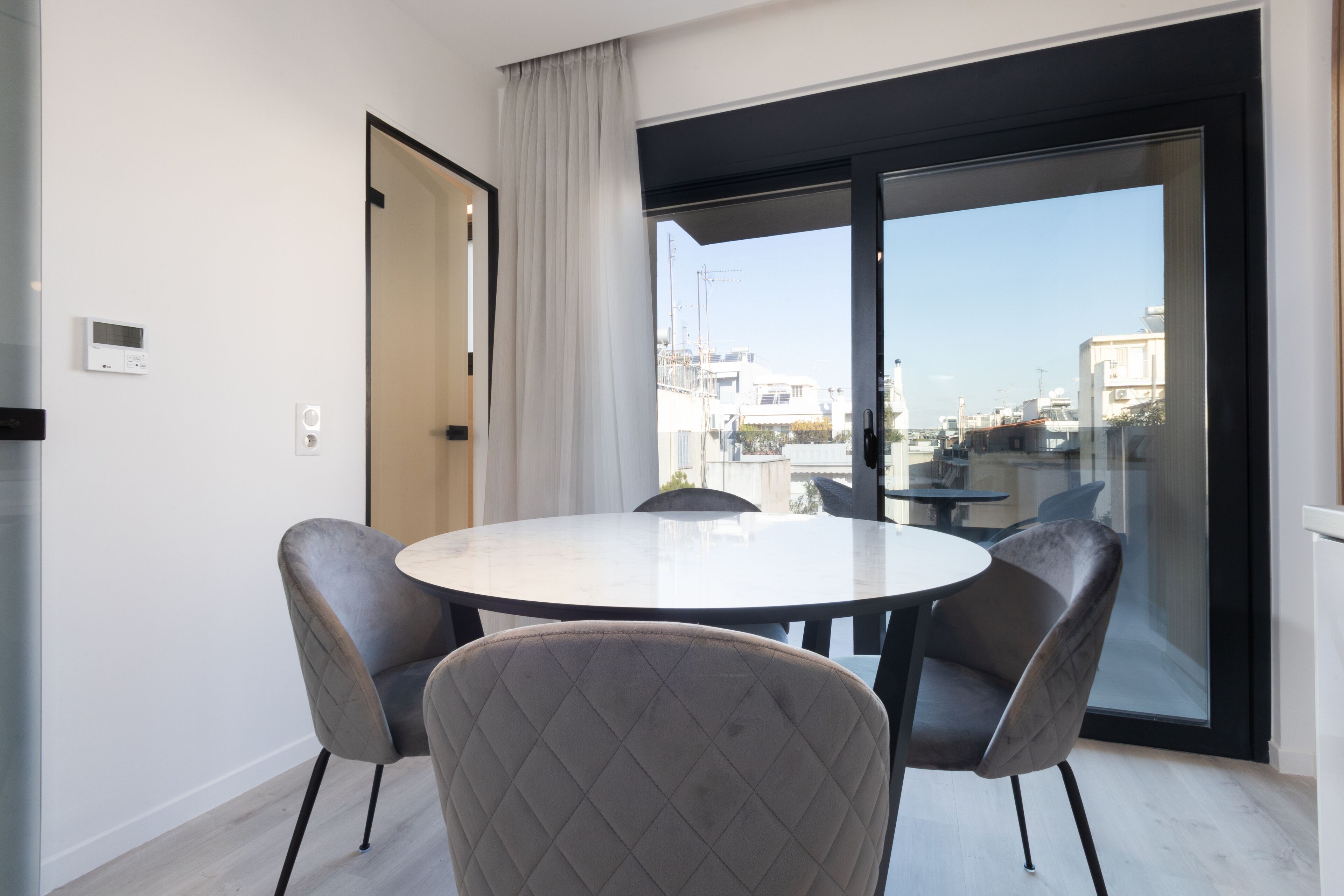 A modern dining area with a round table and four chairs, a sliding glass door leading to an outdoor balcony, and a view of the cityscape.