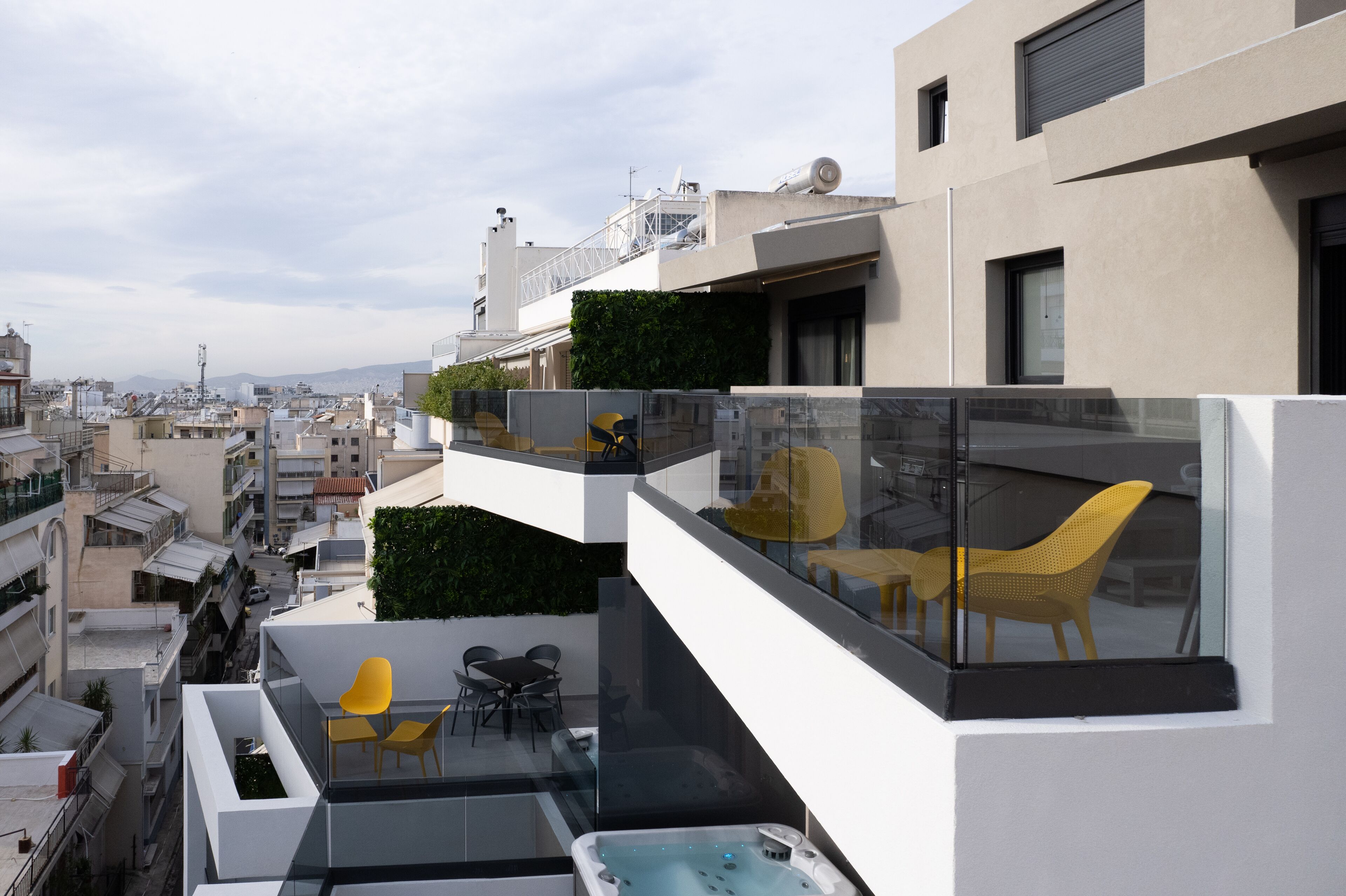 A modern building with a rooftop terrace featuring a hot tub, yellow chairs, and a glass balustrade.