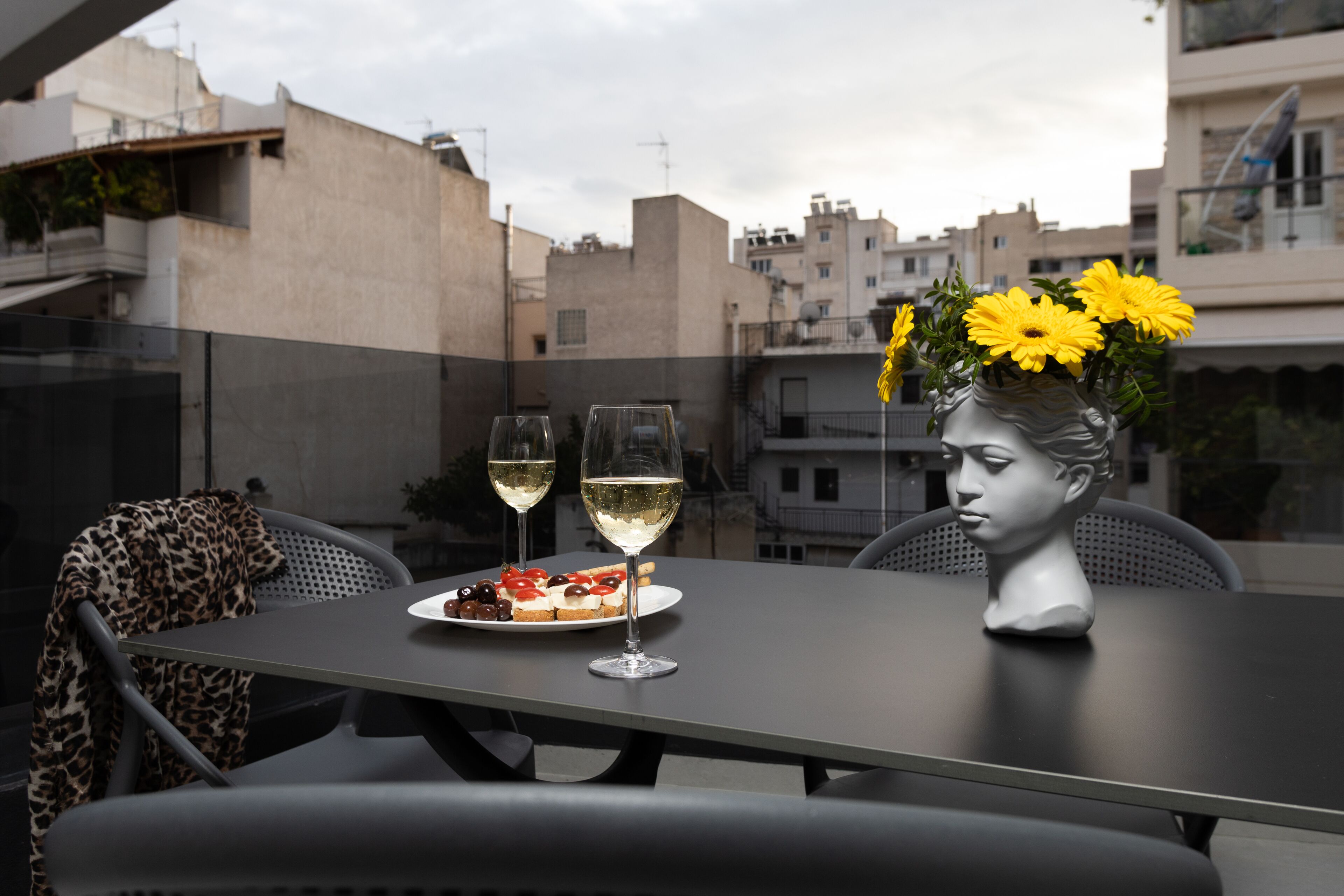 A table with two glasses of white wine, a plate with food, and a vase with yellow flowers on a balcony overlooking a cityscape.