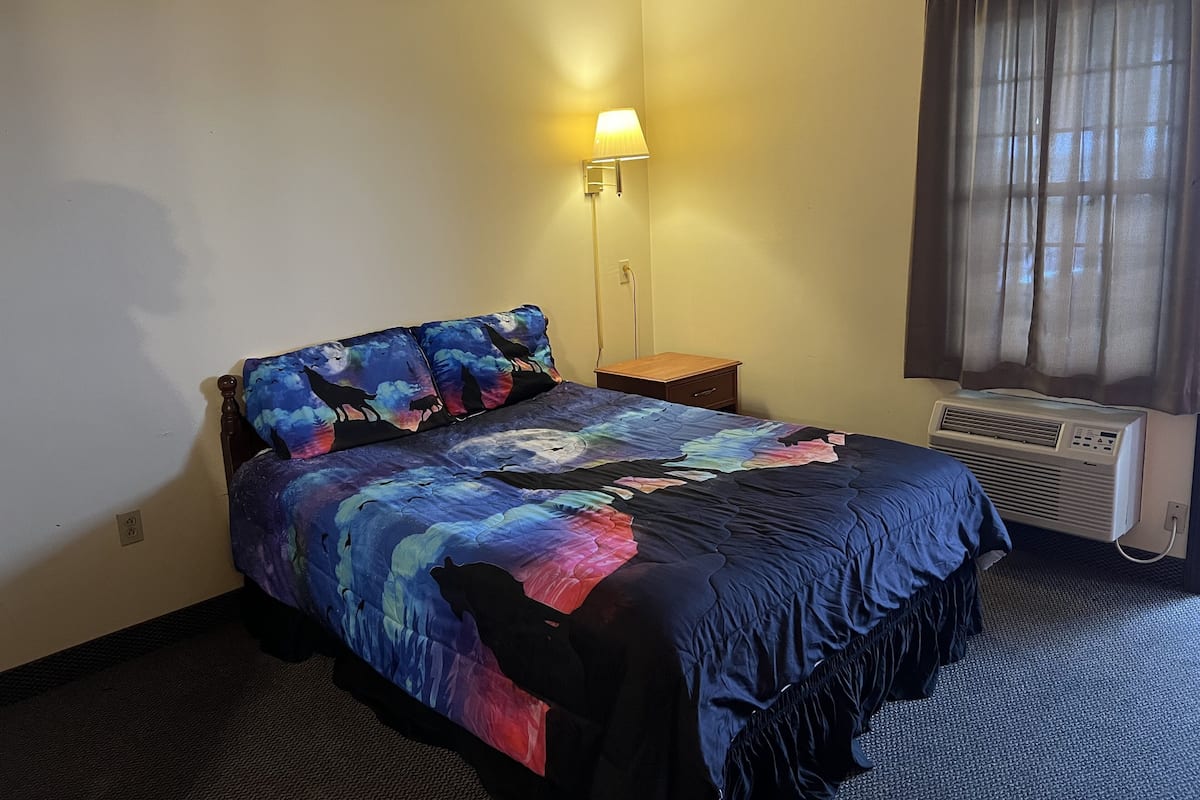 A hotel room with a bed, a nightstand, a lamp, and an air conditioning unit.