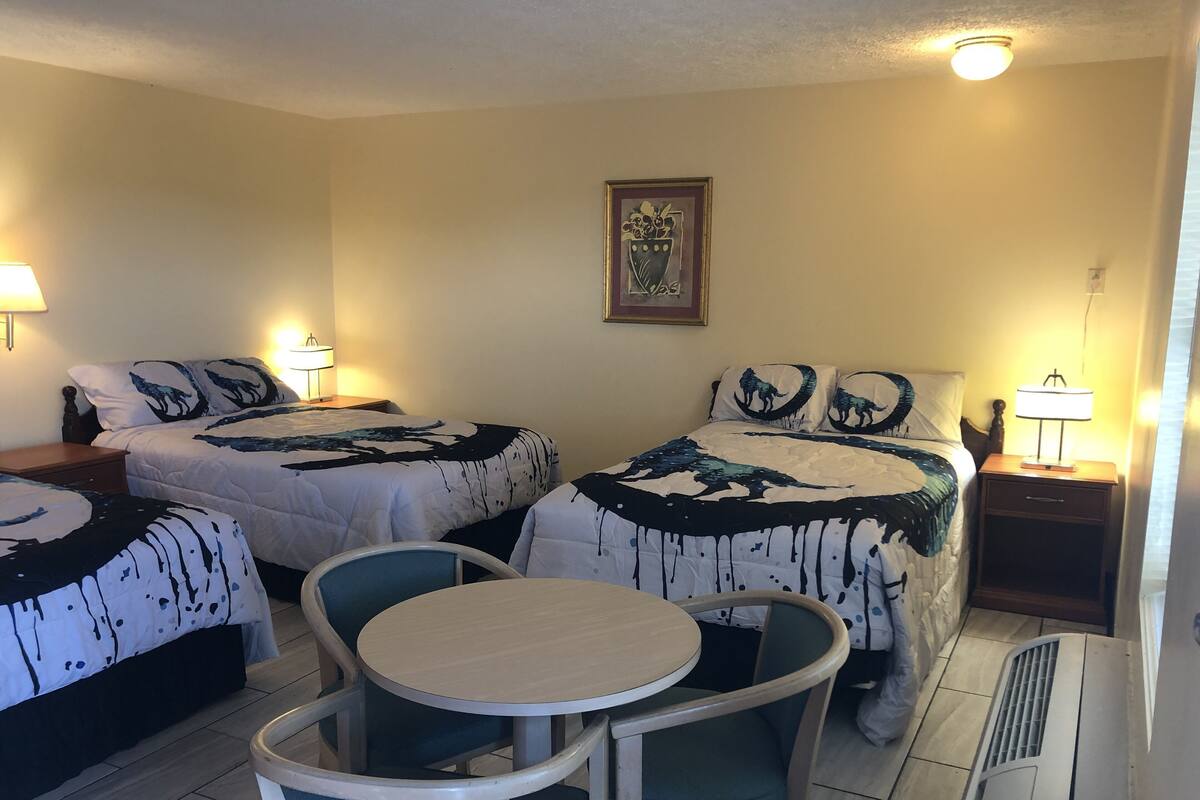 A hotel room with two beds, a round table, and chairs. There are bedside lamps and a ceiling light.