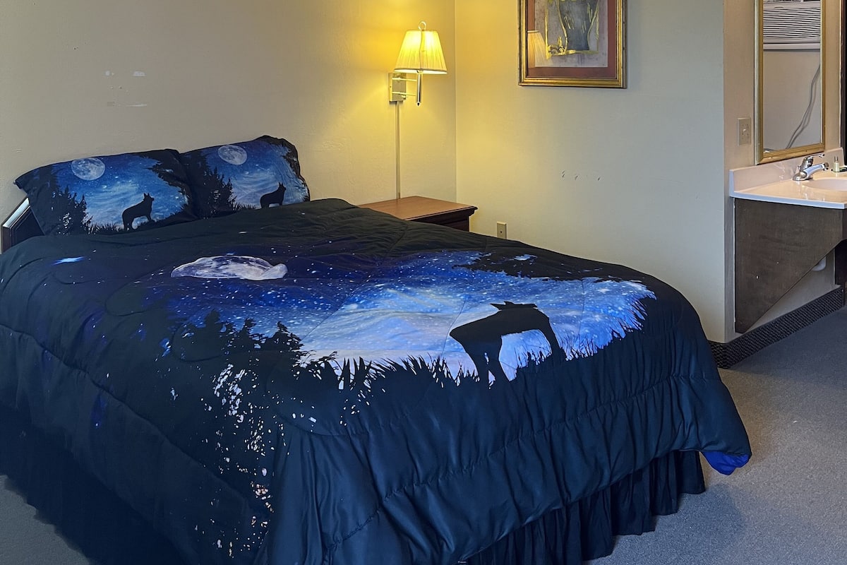 A hotel room with a bed, a nightstand, a lamp, a painting, and a bathroom mirror.
