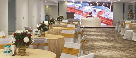 Un salón de banquetes con mesas redondas, sillas blancas con cintas doradas y una pantalla grande que muestra el interior de un restaurante.