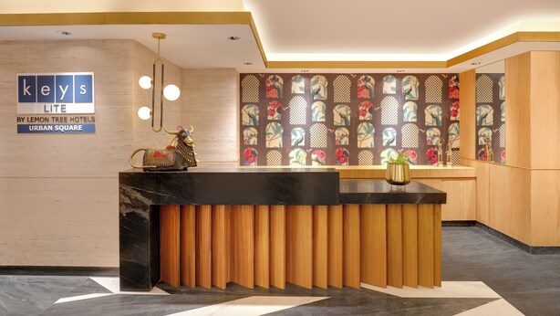 A hotel reception desk with a black marble top, wooden paneling, and a decorative vase. A sign above the desk reads "keys LITE BY LEMON TREE HOTELS URBAN SQUARE." - Keys Lite by Lemon Tree Hotels, Urban Square, Udaipur (Udaipur)