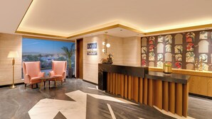 A hotel reception area with a reception desk, two pink chairs, a lamp, and a view of the cityscape.