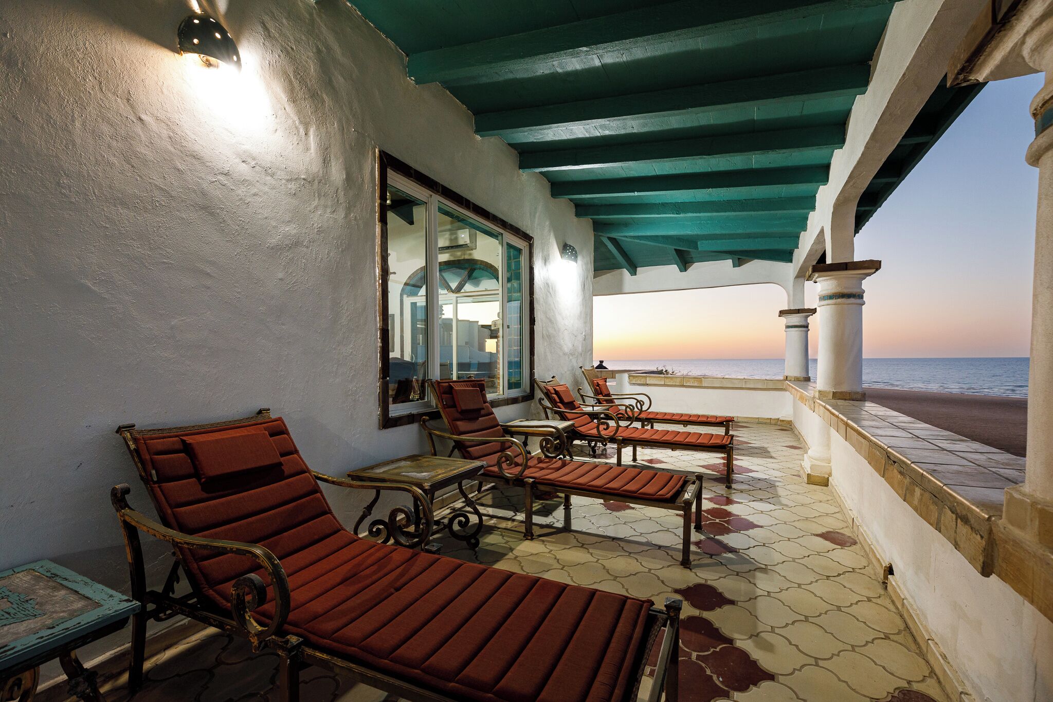 A covered outdoor seating area with lounge chairs and a table, overlooking a body of water.
