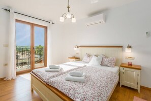 A bedroom with a large bed, a nightstand, a wooden headboard, a ceiling light, and a view of the outdoors.