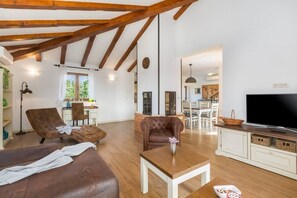 A spacious living area with a wooden ceiling, leather sofas, a TV, and a dining area in the background.