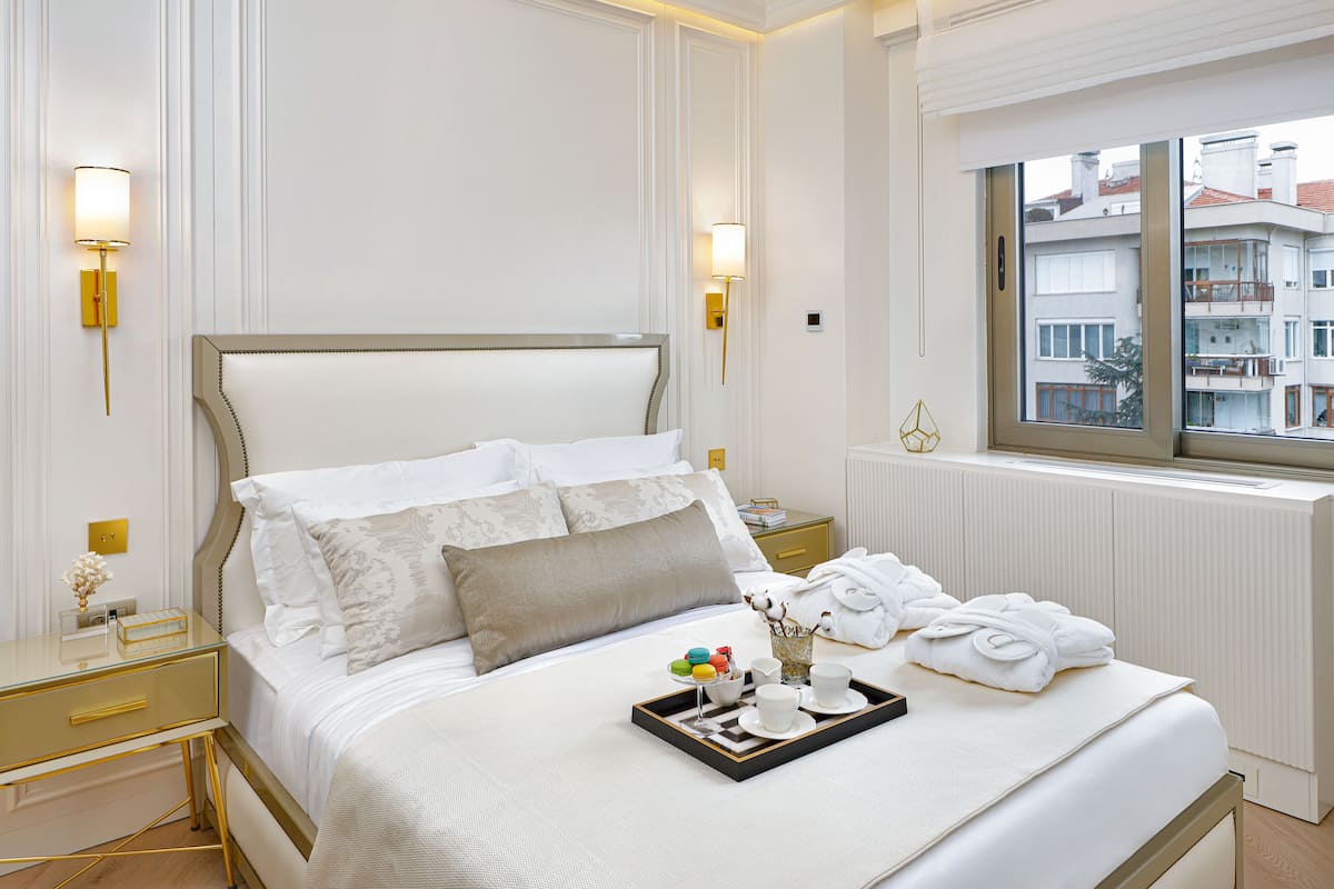 Un lit bien fait, avec du linge de lit blanc, une table de chevet avec une lampe et une fenêtre donnant sur des immeubles.