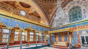 Een rijk gedecoreerde kamer met gedetailleerde blauwe en gouden patronen, een kroonluchter en een groot raam.