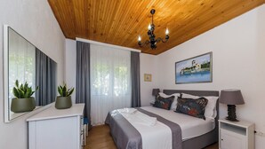 Ein Schlafzimmer mit Holzboden, einem Bett mit grau-weißer Bettdecke, einem Nachttisch mit Lampe, einer kleinen Kommode und einem Bild an der Wand.