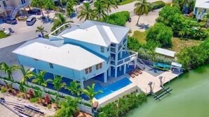 Un edificio grande de dos pisos con piscina y muelle, rodeado de vegetación exuberante y palmeras.