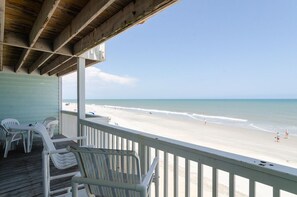 A beach view from a covered deck with white plastic chairs and a wooden railing. - Spacious ocean front condo in Ocean Dunes! (Kure Beach)
