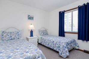 A bedroom with two single beds, a nightstand with a lamp, a window with blue curtains, and a framed picture on the wall. - Spacious ocean front condo in Ocean Dunes! (Kure Beach)